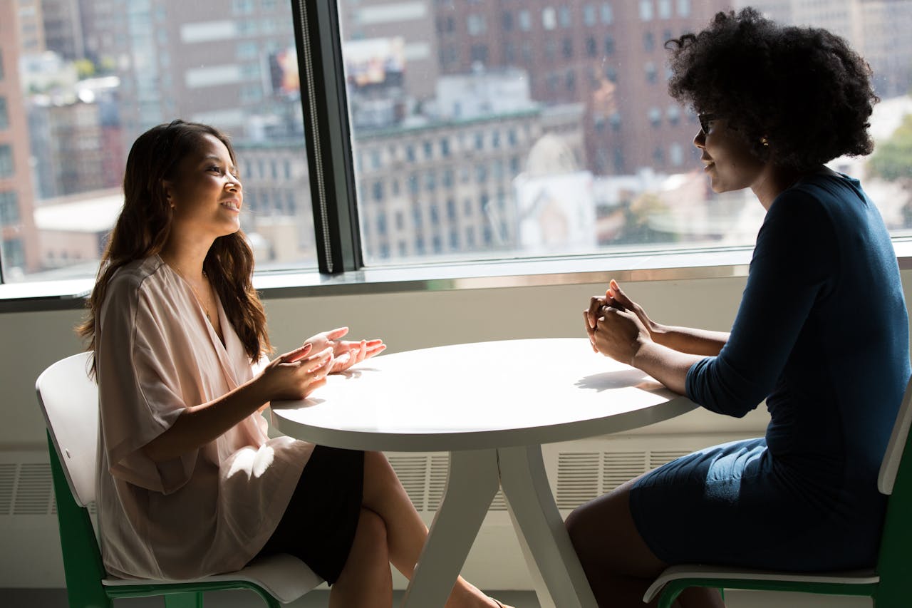 who-we-are Two professional women having a discussion at a desk in a bright office setting.